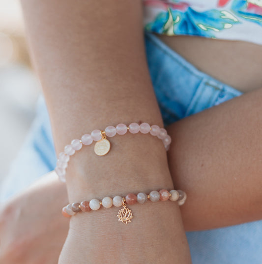 Poignet de femme portant deux bracelets superposés en pierres naturelles : bracelet en Quartz Rose avec médaille dorée chakra Anahata et bracelet en Pierre de Lune avec breloque Fleur de Lotus dorée. Bijoux spirituels Andréa Chereau.