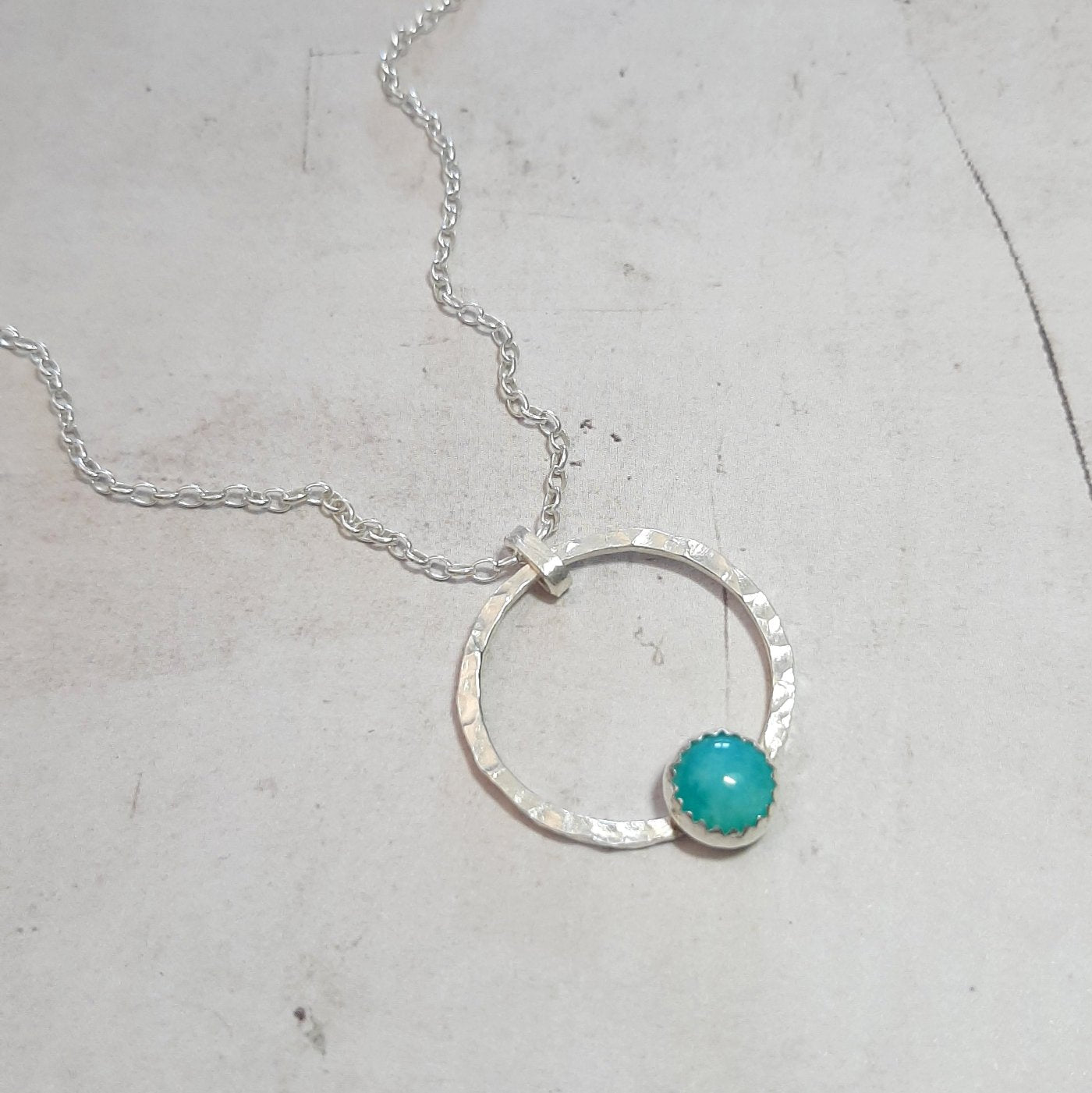 Le collier en argent massif se distingue par une chaîne brillante et un pendentif rond à l’effet martelé, magnifiquement orné d’une pierre d’amazonite sertie. L'élégance intemporelle du design allie parfaitement modernité et tradition, faisant de ce bijou un choix idéal pour toutes les occasions, qu'il s'agisse d'une tenue décontractée ou d'une soirée habillée. L'amazonite, connue pour ses vertus apaisantes, ajoute une touche de couleur et d'énergie positive, renforçant l'attrait de ce bijou exceptionnel.