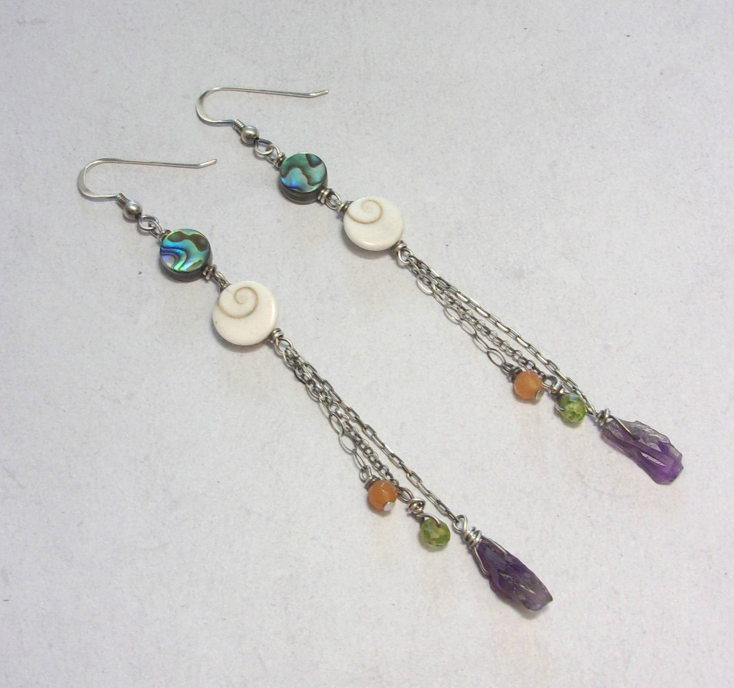 Boucles d'oreilles ethniques longues en argent patiné noir, ornées de coquillages et de pierres naturelles telles que l'améthyste, le péridot et l'aventurine. Les pierres et coquillages, étant naturels, présentent des variations de forme et de couleur. La longueur est de 10 cm, offrant un design unique et élégant.