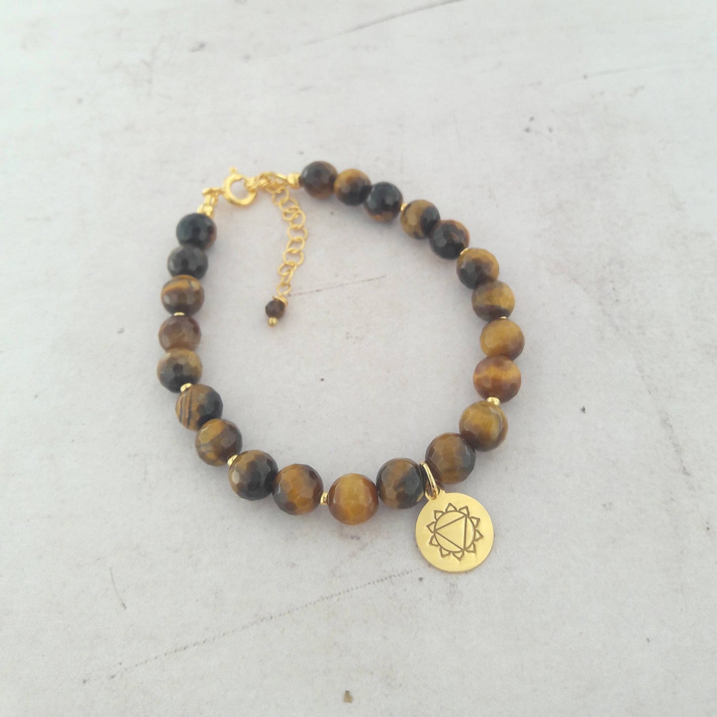 Bracelet en œil de tigre et argent 925 doré, orné d’une médaille représentant le chakra Manipura, lié au plexus solaire. Longueur ajustable de 17 cm avec chaînette. L’œil de tigre, pierre protectrice, renvoie les énergies négatives tout en apportant vitalité et confiance en soi. Le chakra Manipura symbolise la puissance personnelle et l’estime de soi, favorisant l’ouverture aux autres et une attitude positive.