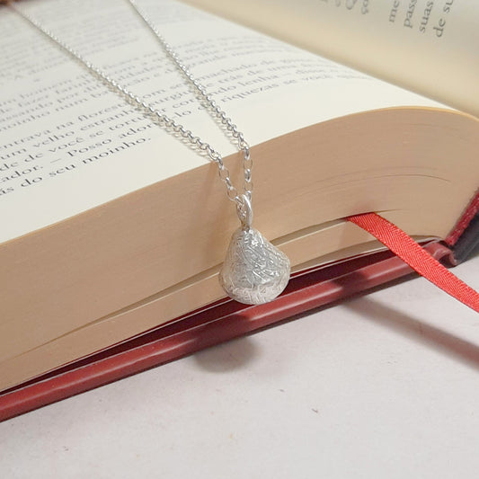 Collier appeleur d’anges en argent 925, pendentif grelot texturé et chaîne posés sur un livre ouvert.