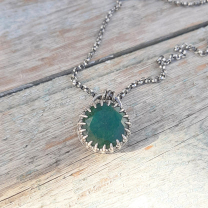 Gros plan du pendentif en émeraude verte, entouré de griffes argentées, sur un support en bois.
