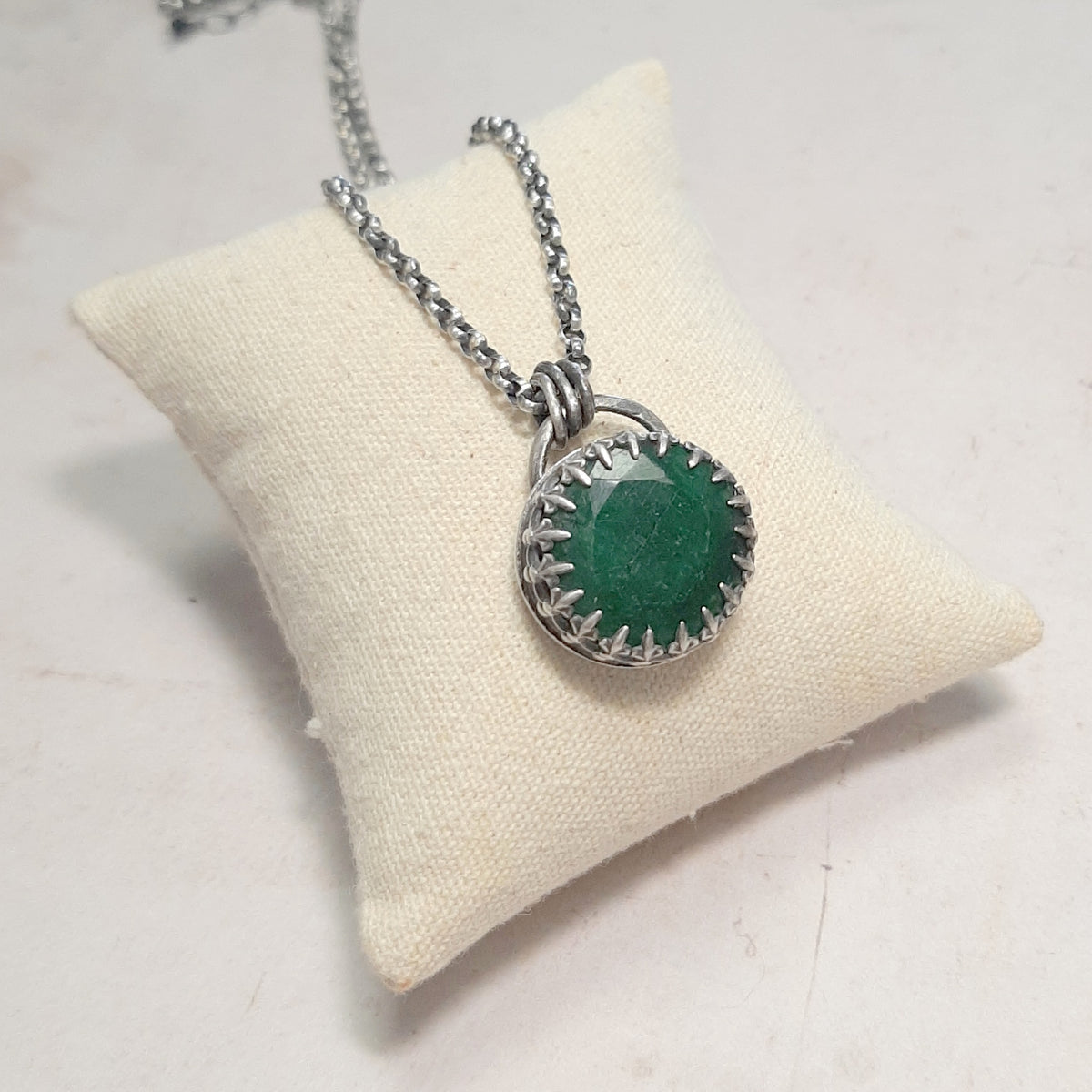  Pendentif en émeraude facettée avec monture  à griffes en argent patiné, présenté sur un coussin écru.