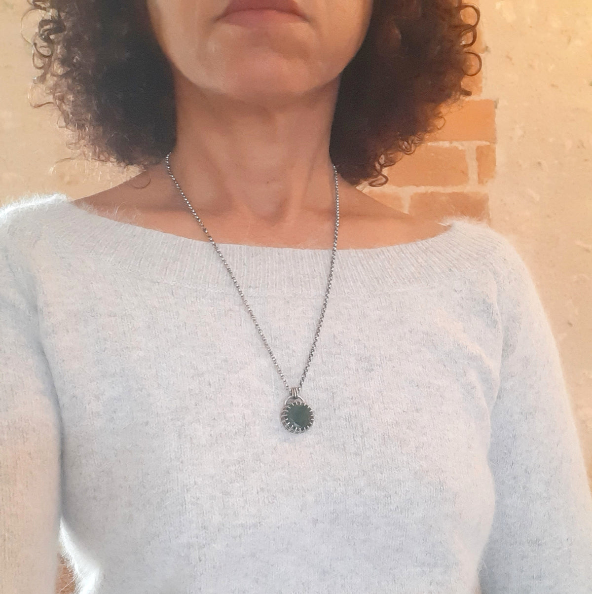 Collier avec pendentif rond en émeraude porté sur un pull clair, montrant sa longueur sur le buste.