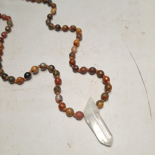 Mala 108 perles en jaspe Picasso tons rouges, verts et ocres avec pendentif en cristal de roche, disposé sur fond clair 