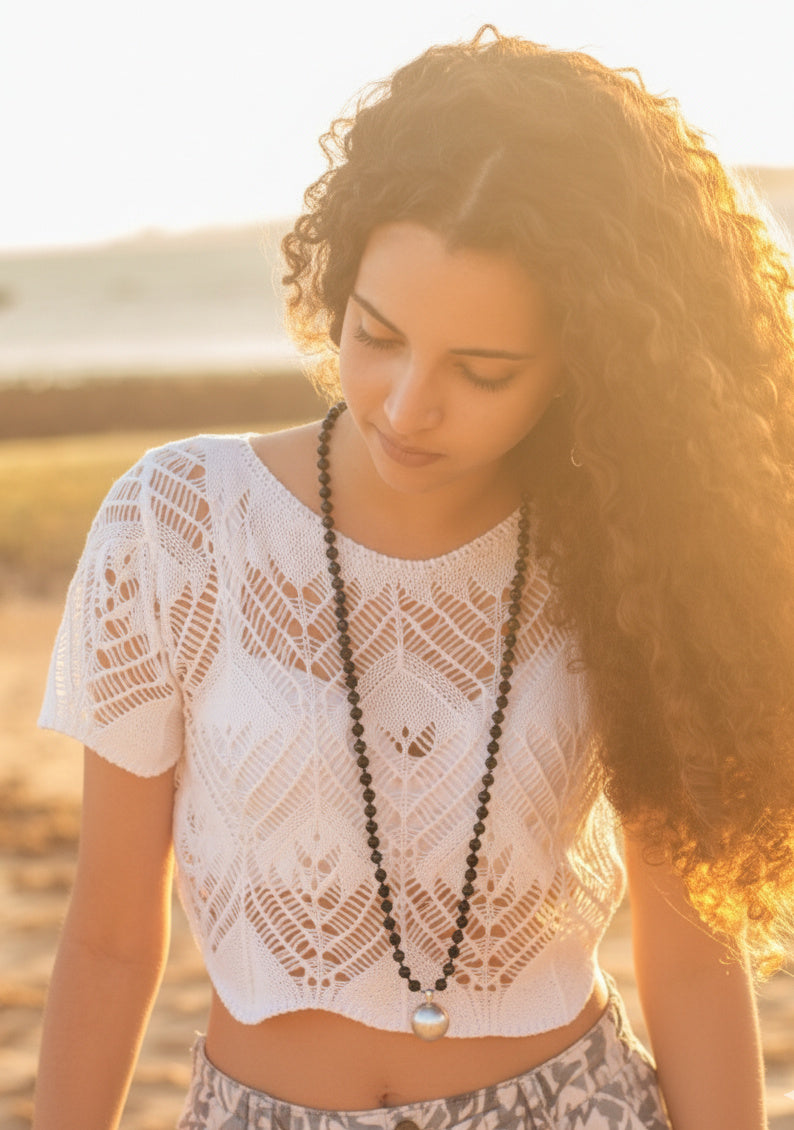 Ambiance dorée d'un coucher de soleil sur la plage, mettant en valeur une femme portant un élégant sautoir mala de perles noires en onyx et un pendentif argenté.