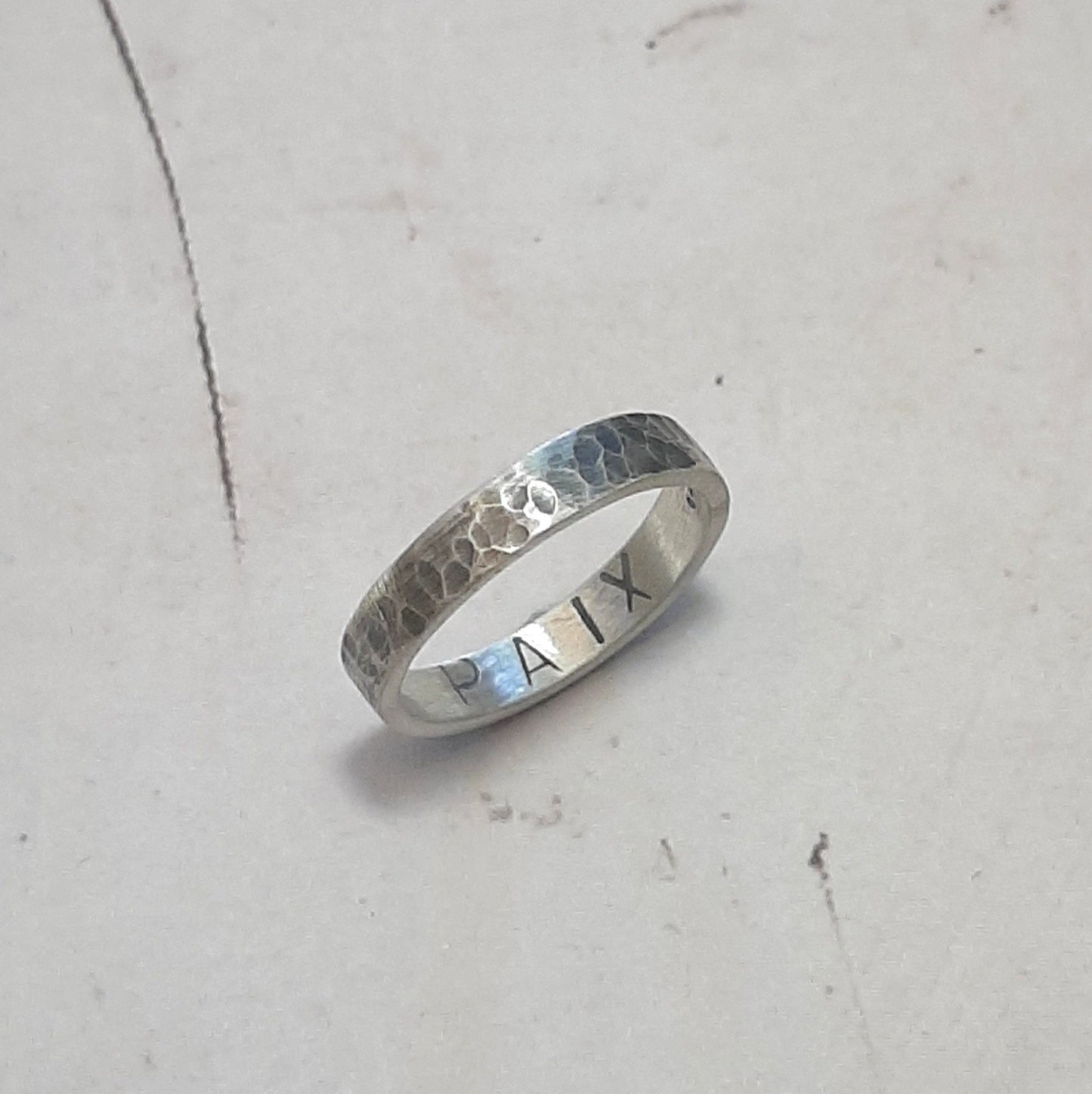 Bague en argent massif martelé et patiné, au design unique et artisanal. L'intérieur de l'anneau présente une gravure personnalisable, jusqu'à 6 caractères, offrant la possibilité d'y inscrire un message, une date ou un prénom. Avec une largeur d'environ 4 mm, cette bague allie élégance et sobriété. Les demandes spécifiques pour une création sur mesure sont les bienvenues.
