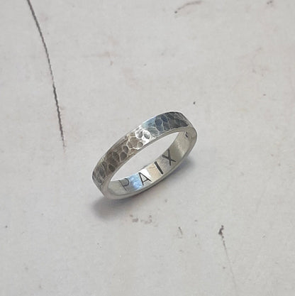 Bague en argent massif martelé et patiné, au design unique et artisanal. L'intérieur de l'anneau présente une gravure personnalisable, jusqu'à 6 caractères, offrant la possibilité d'y inscrire un message, une date ou un prénom. Avec une largeur d'environ 4 mm, cette bague allie élégance et sobriété. Les demandes spécifiques pour une création sur mesure sont les bienvenues.