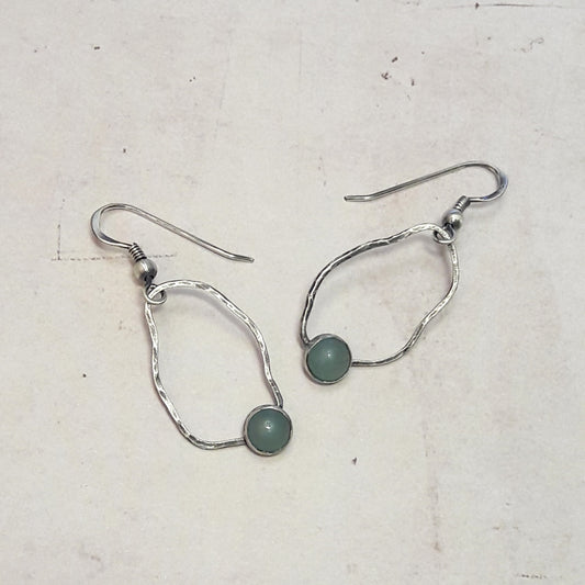 Boucles d'oreilles ethniques en argent massif patiné, ornées de pierres naturelles d'amazonite. Chaque pièce présente un cercle irrégulier et texturé, mettant en valeur une pierre d'amazonite de 6 mm de diamètre. La longueur totale est de 4 cm. L'amazonite, pierre apaisante, éveille la joie de vivre et aide à vivre l'instant présent.