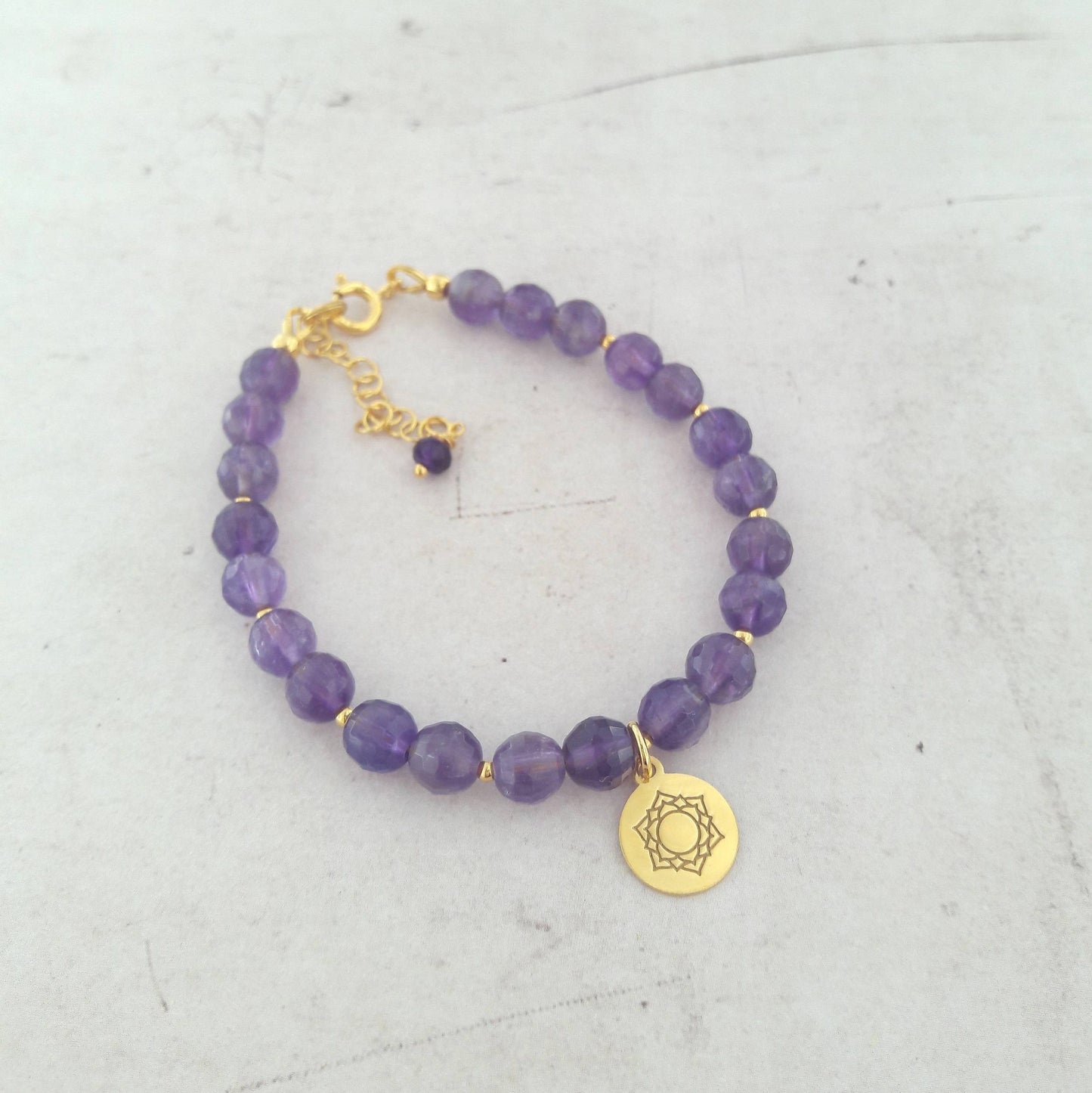 Bracelet en pierres naturelles d’améthyste avec une médaille représentant le chakra Sahasrara, en argent 925 doré. Longueur ajustable de 17 cm, avec une chaînette. L’améthyste, d’un diamètre de 6 mm, est reconnue pour ses vertus apaisantes, associée à la sérénité et à la sagesse. Reconnue pour renforcer l’intuition et favoriser la méditation, cette pierre soutient l’équilibre émotionnel tout en étant un symbole de force intérieure.